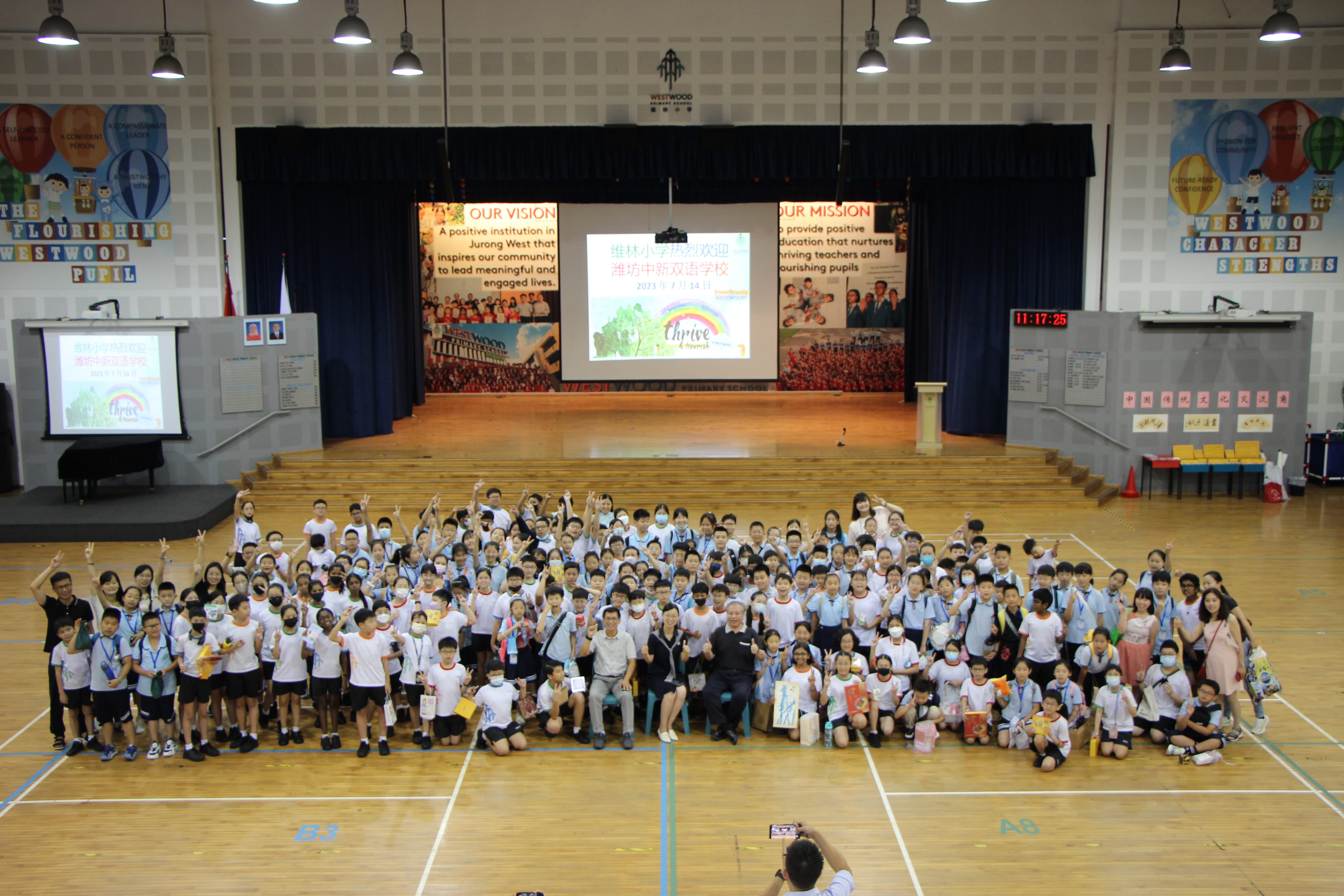 Large group of students and teachers posing in Westwood Primary School hall with a projected welcome screen on stage.